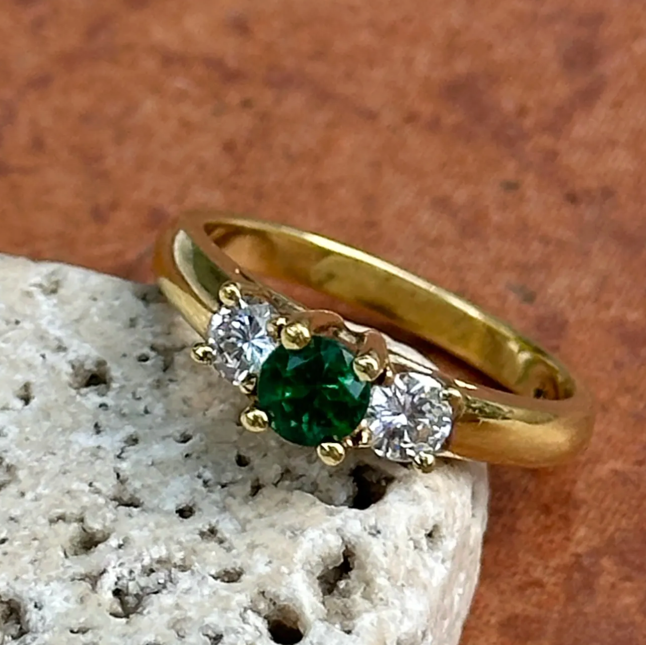 A yellow gold three-stone ring featuring a round green emerald center stone and two round clear accent stones, displayed on a textured light stone against a warm brown background.