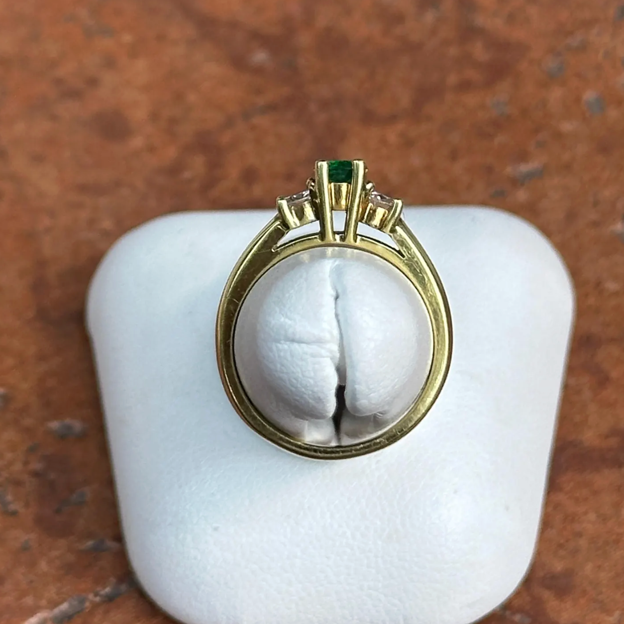 A vintage-style gold ring with a green emerald-cut center stone and small clear stones, displayed on a white ring stand against a brown textured background.