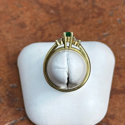 A vintage-style gold ring with a green emerald-cut center stone and small clear stones, displayed on a white ring stand against a brown textured background.